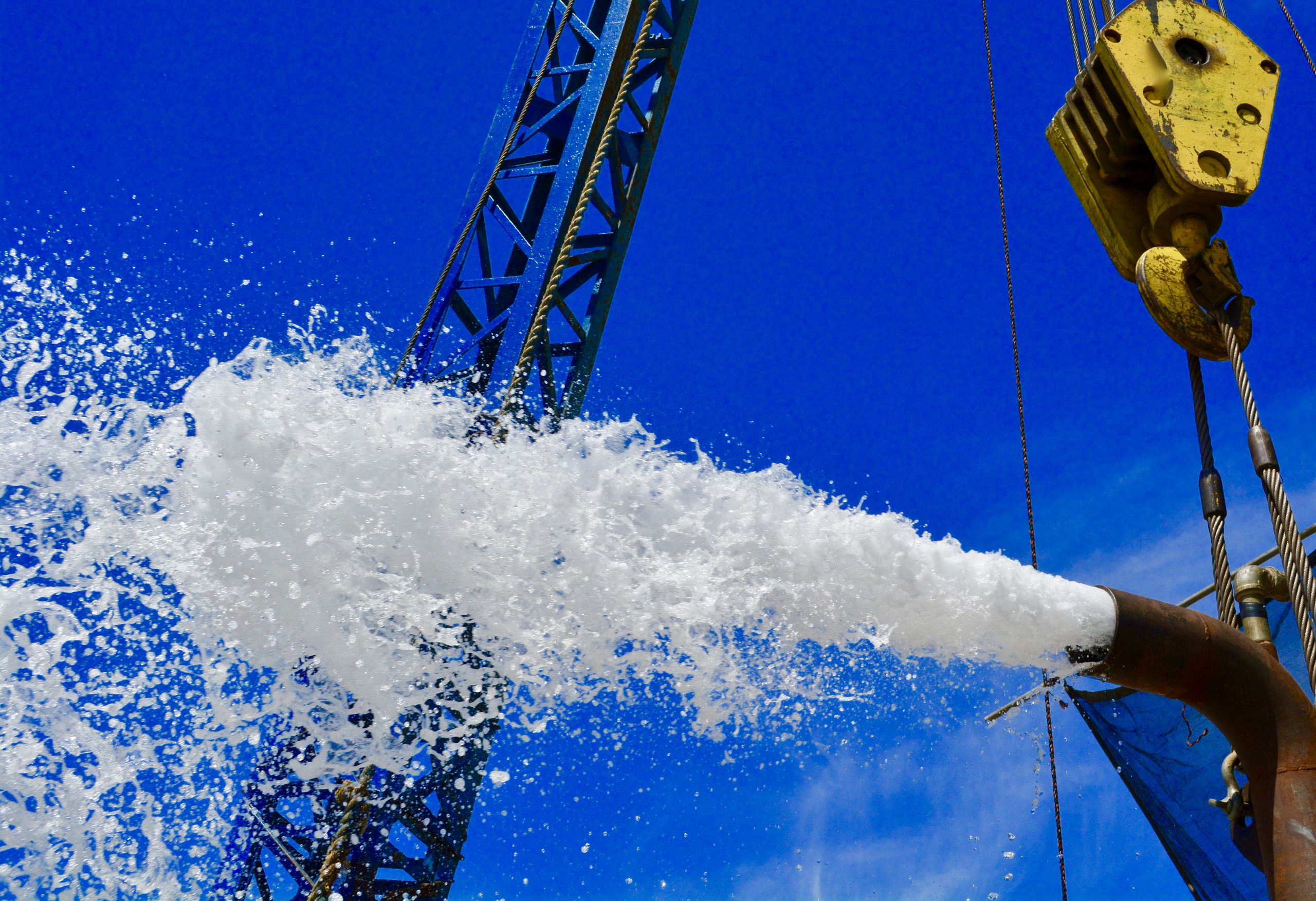 Water and air blasting from a discharge pipe during air-lift well development, drill rig derrick in the background.