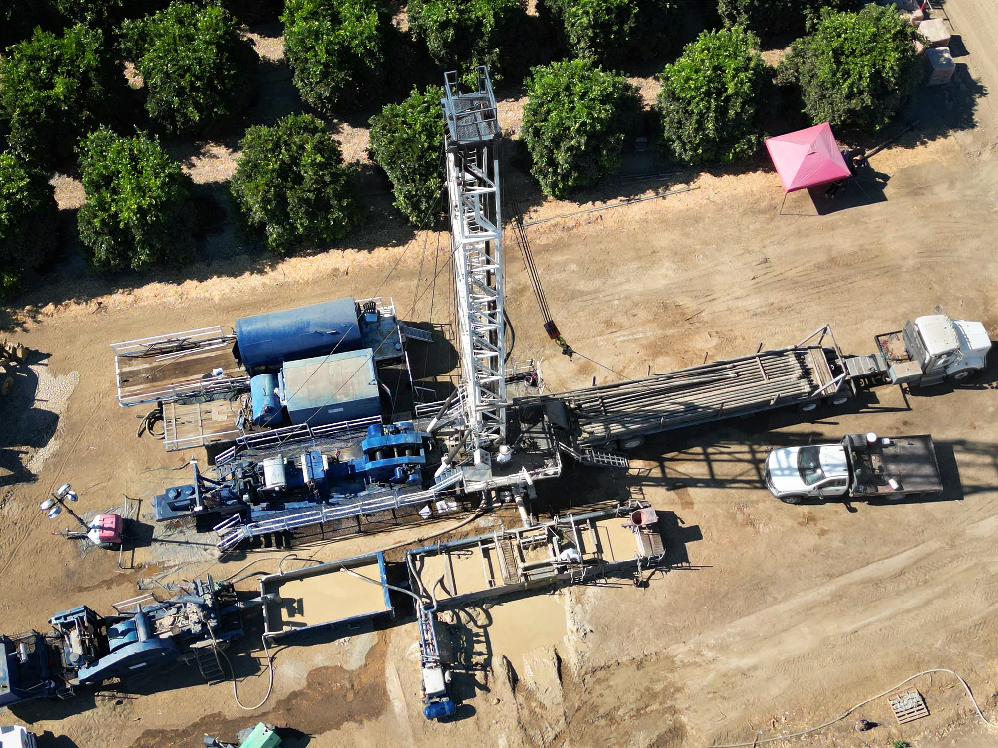 Aerial view of a water well drilling rig set up beside an orchard, with support trucks and mountains in the background.